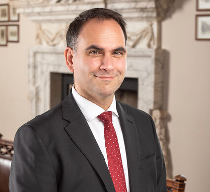 A smiling man in a black suit and red tie standing in a professional setting.