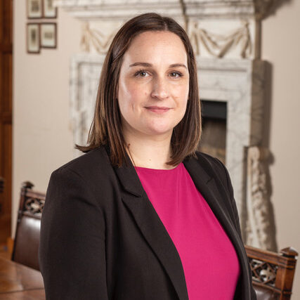 A woman in a professional setting, wearing a black blazer and pink top, standing in front of a fireplace in a formal room.