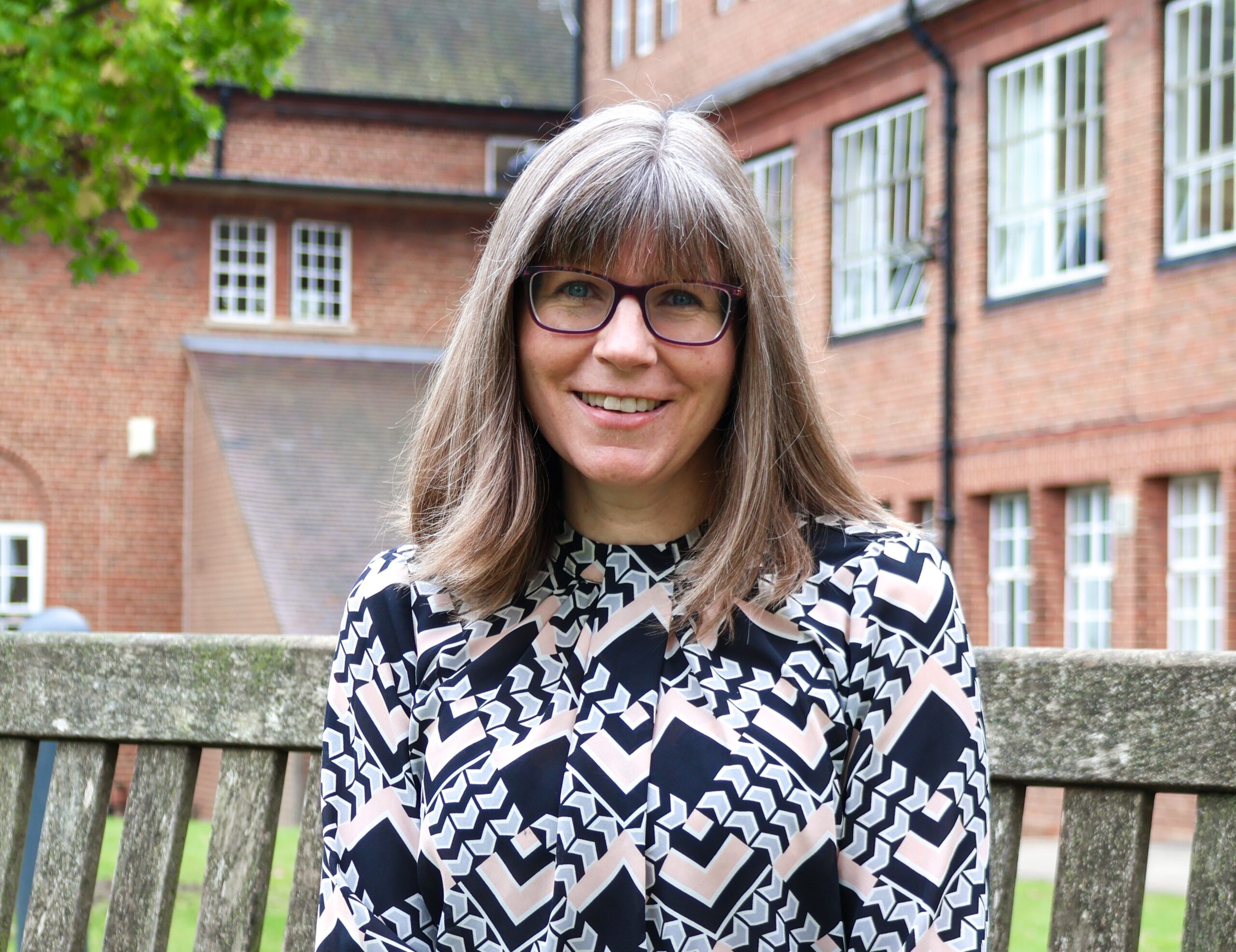 Woman with glasses smiling while sitting on a bench outside a brick building