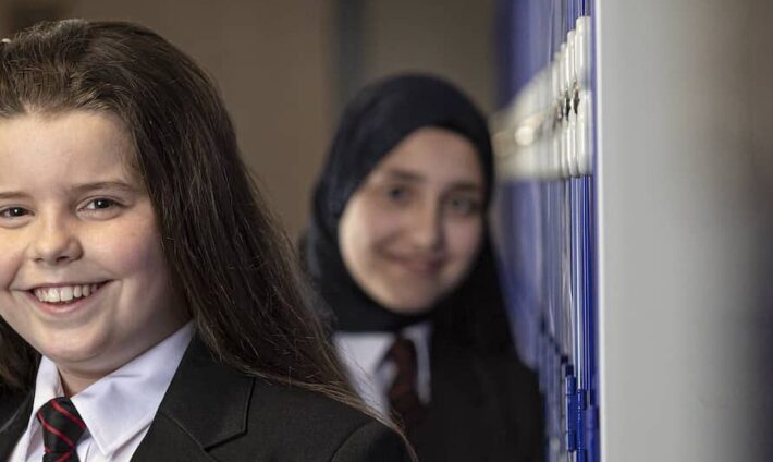 Two students smiling in a school hallway, one wearing a hijab.