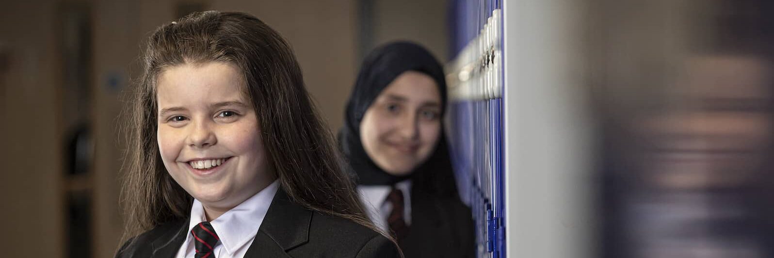 Two students smiling in a school hallway, one wearing a hijab.