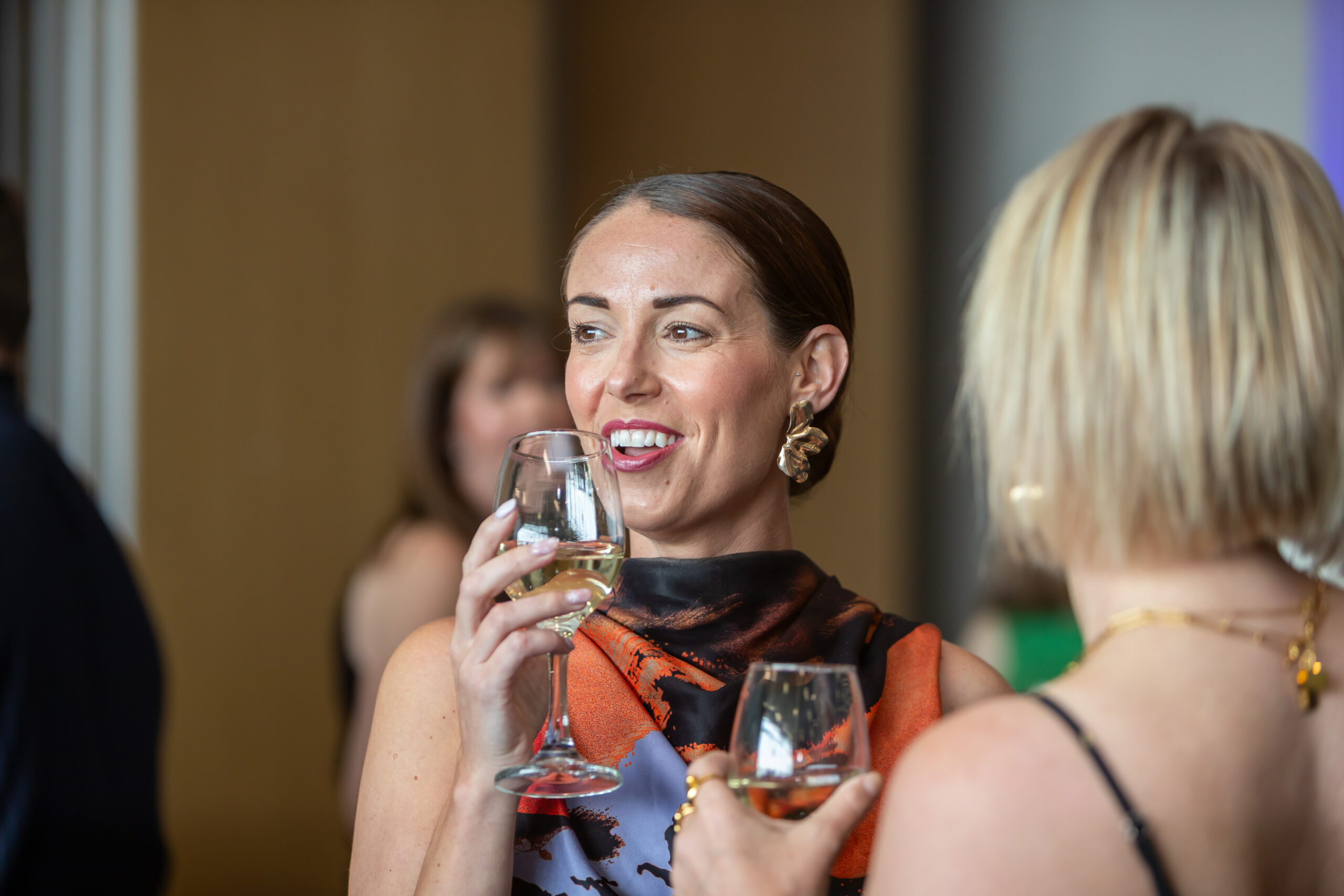 Two women enjoy drinks and conversation smiling together at a social event with people in the background.