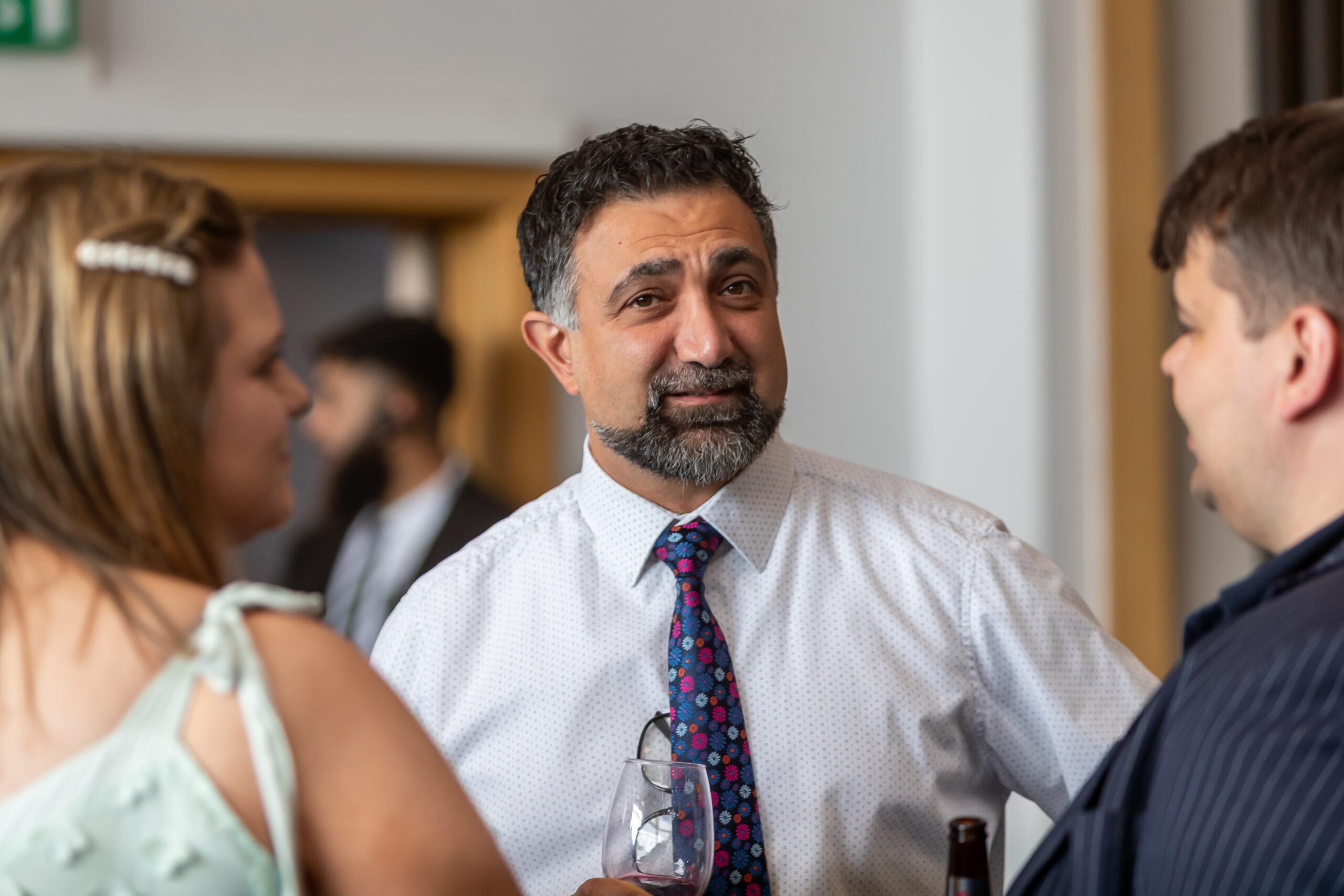 Man with a beard wearing a white shirt and a colourful tie talking to a man and a woman at an event.