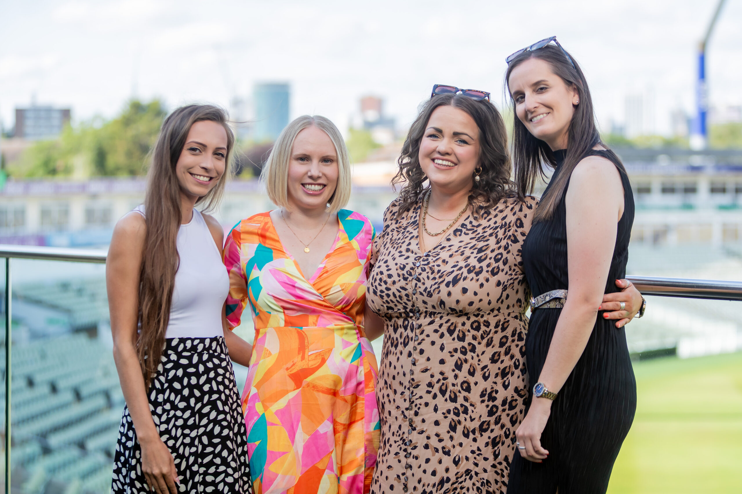 Four smiling women standing together on a balcony overlooking a sports field and city skyline, enjoying a social gathering.