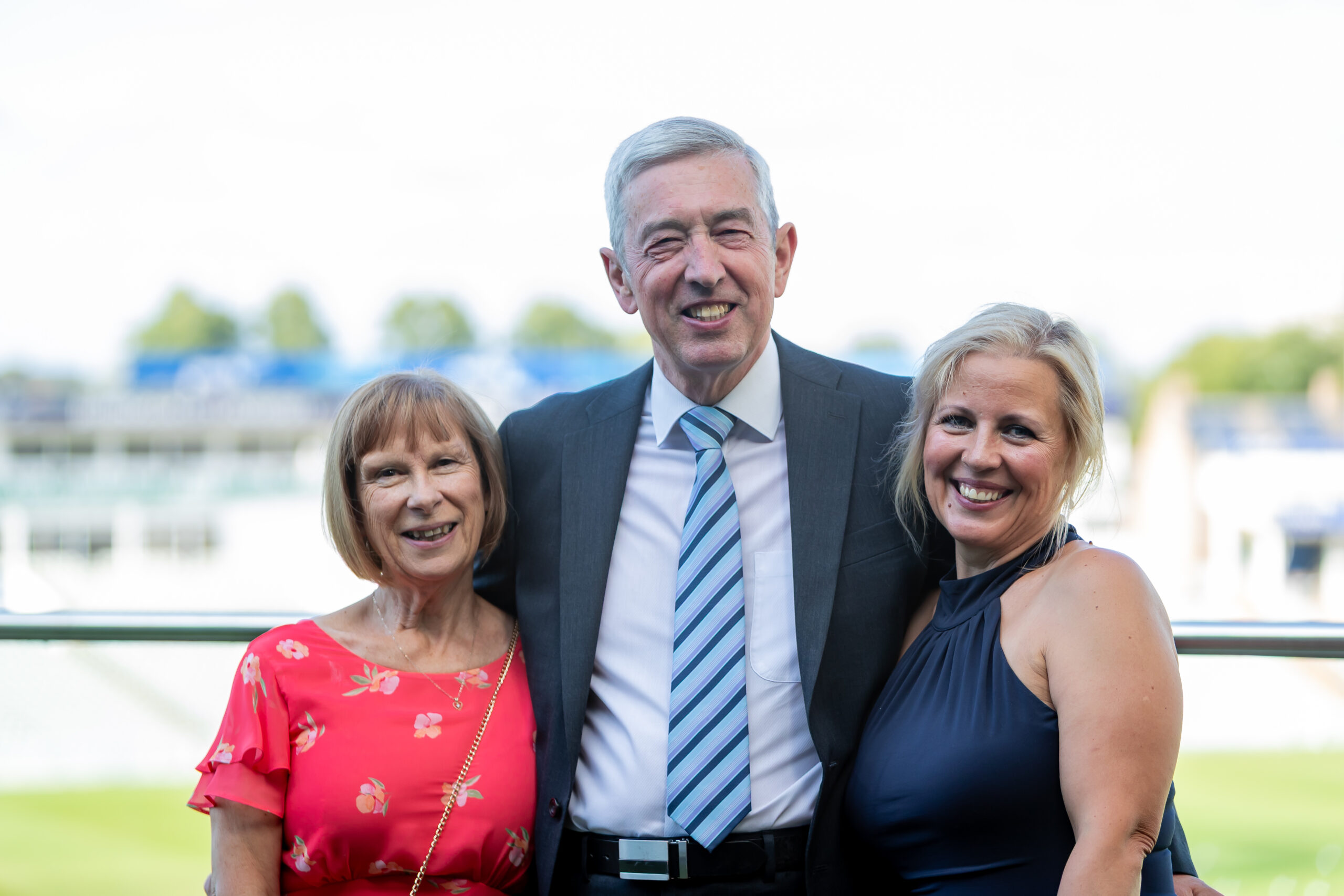A man in a suit standing with two smiling women at a casual outdoor event, likely related to education or community activities.