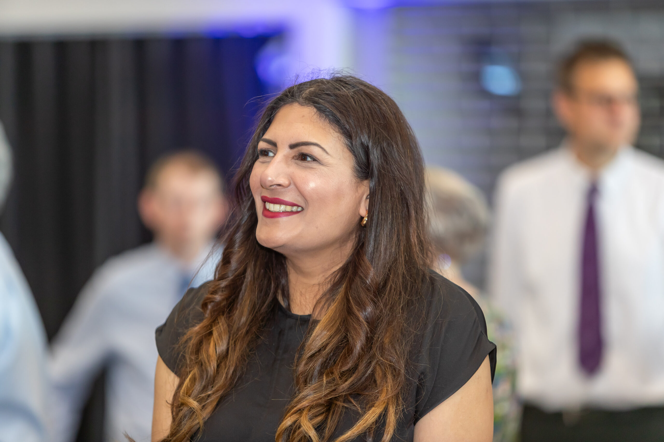 Smiling woman with brunette hair looking to the left amidst a blurred background of people at an educational event.
