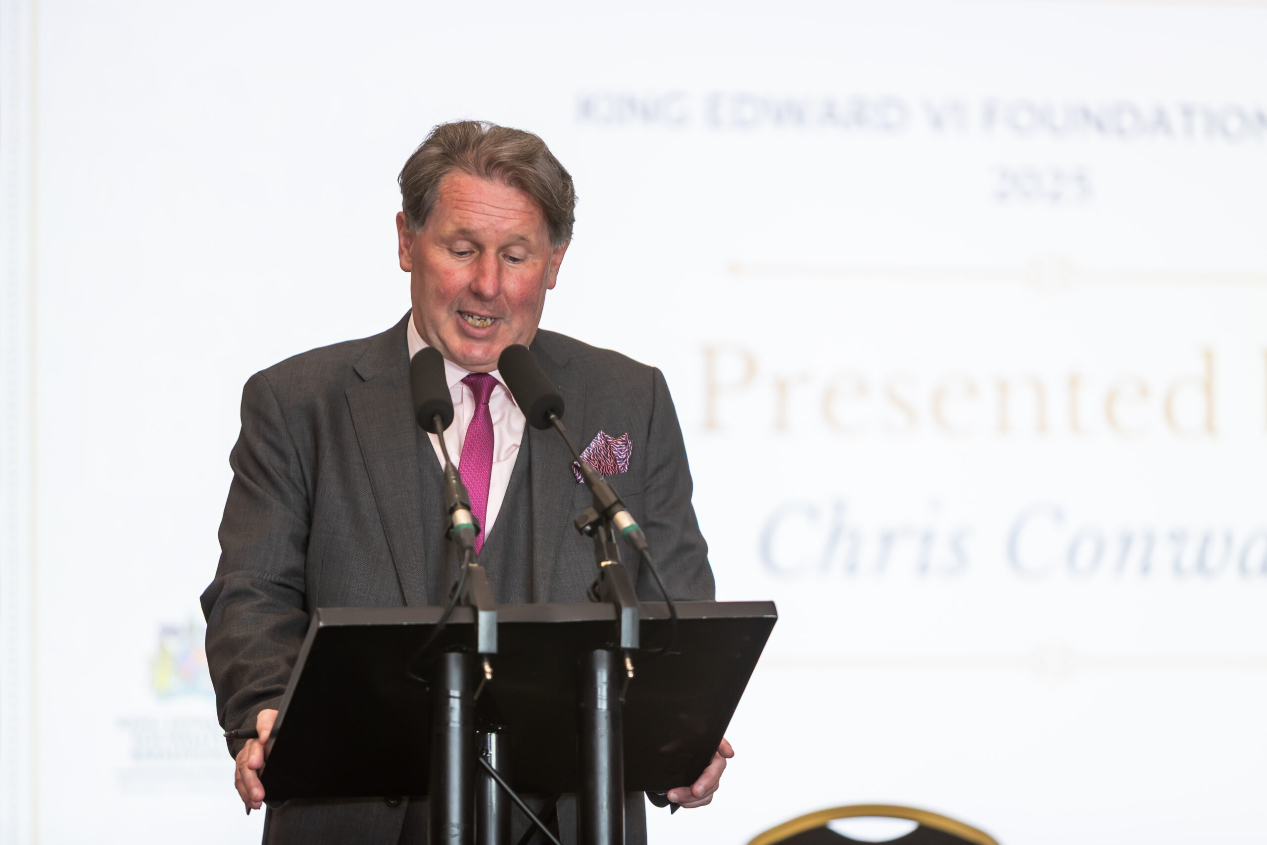 A man in a suit speaking at a podium with two microphones, likely presenting an award at the King Edward VI Foundation.