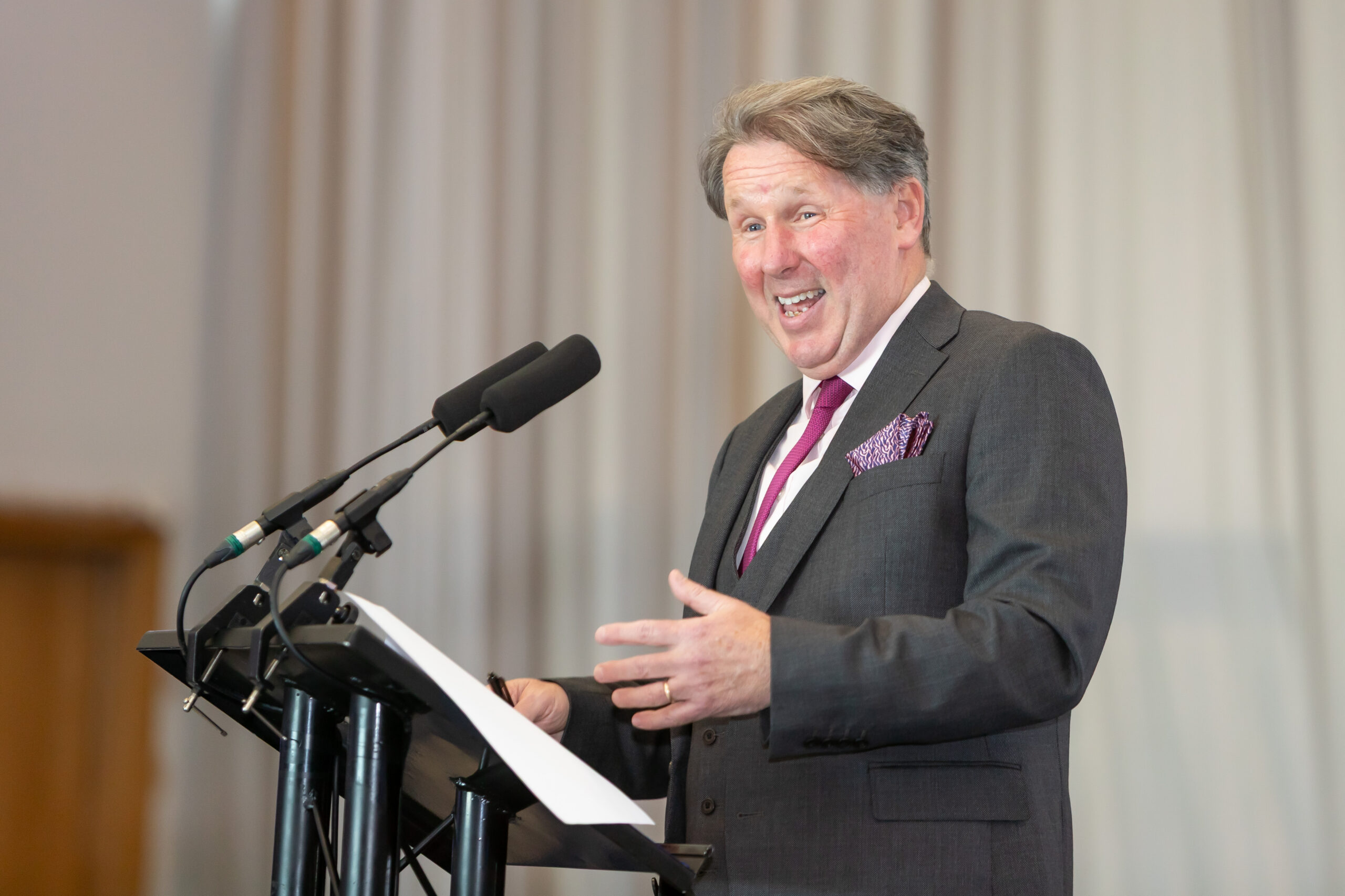 A man in a suit speaking at a lectern with microphones, engaging his audience with a confident gesture.