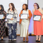 Four diverse students proudly hold certificates on stage, smiling and celebrating their achievement in a formal event, promoting student life and educational accomplishments.