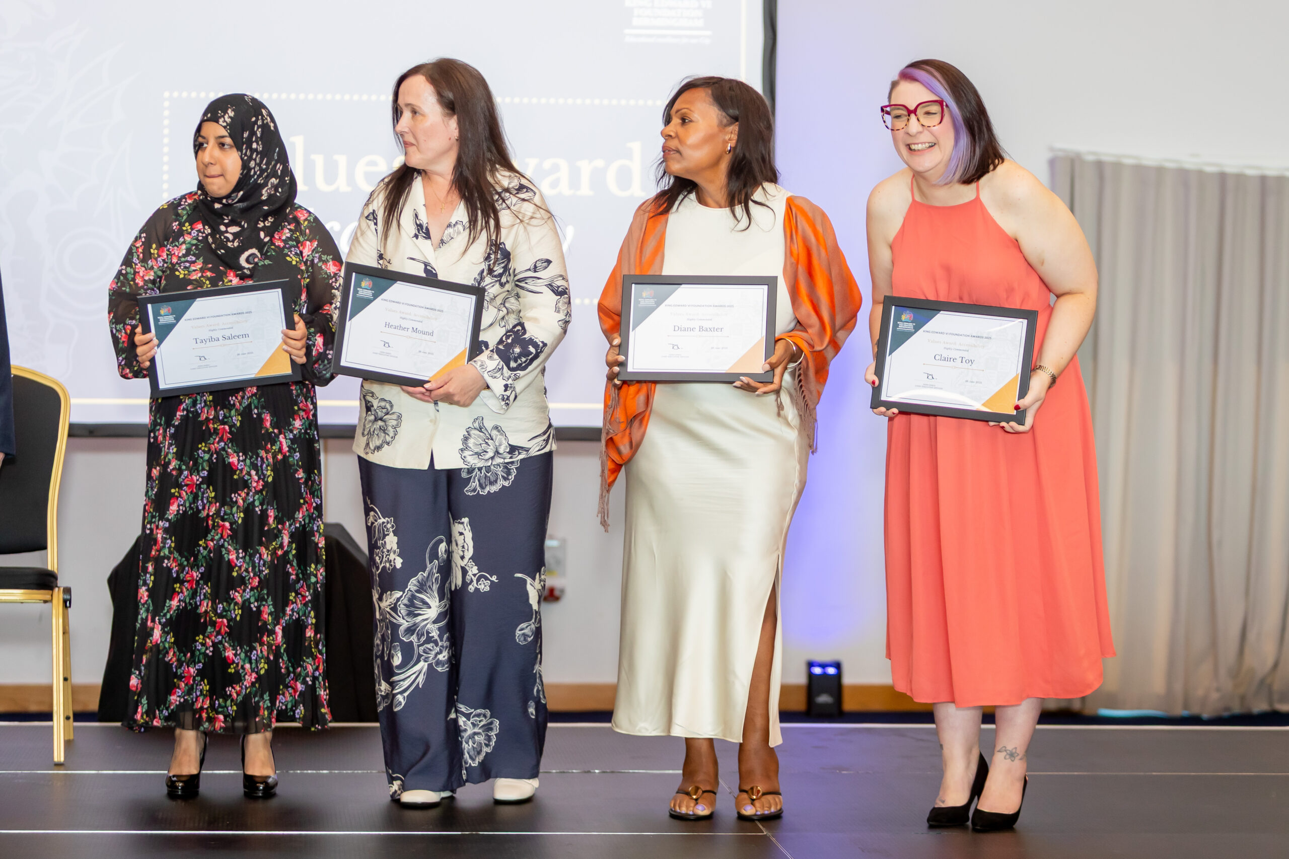 Four diverse students proudly hold certificates on stage, smiling and celebrating their achievement in a formal event, promoting student life and educational accomplishments.