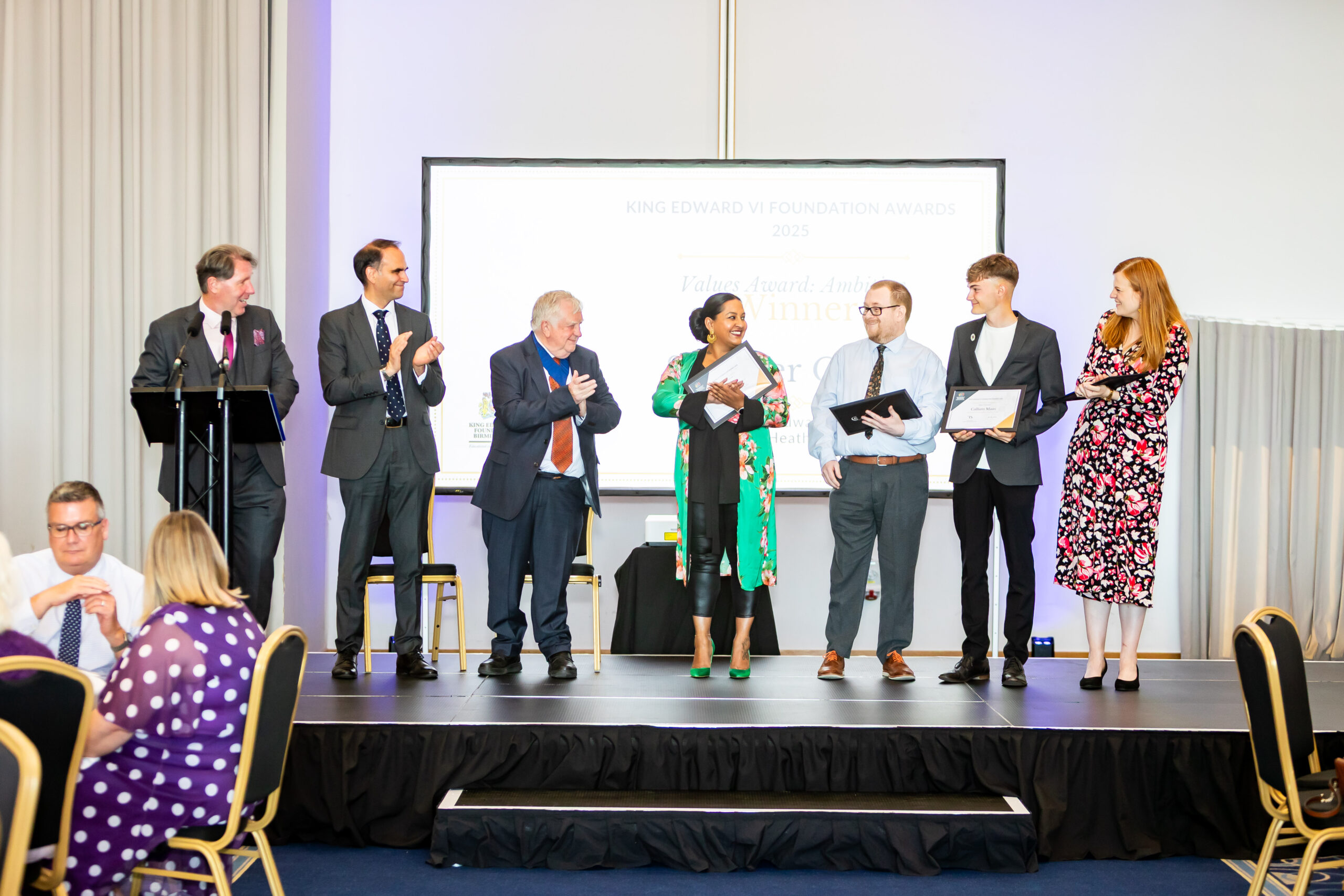 A group of people on stage at the King Edward VI Foundation Awards 2025, clapping and holding certificates, surrounded by an audience seated in gold chairs.