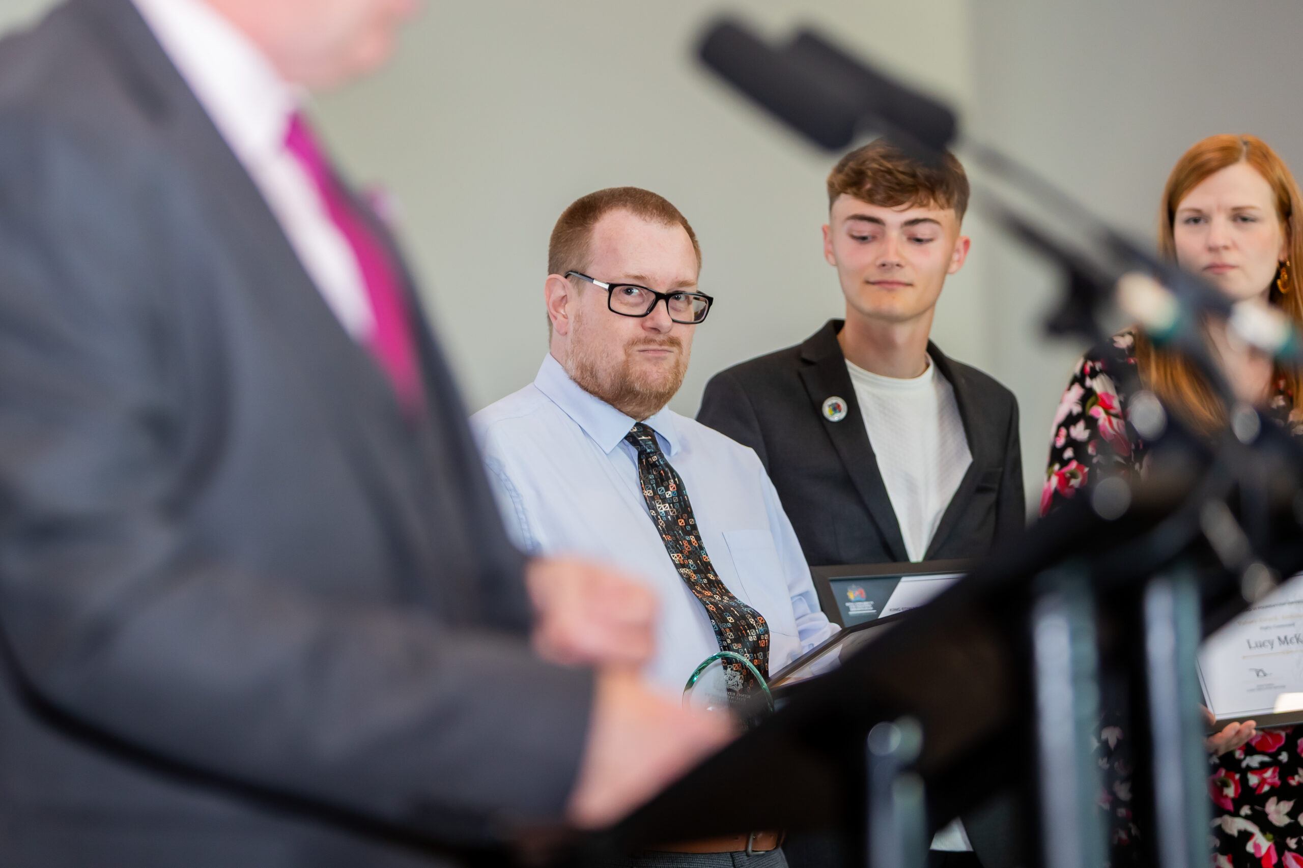 A man in a suit speaking at a podium with three students listening attentively, likely during an educational event or award ceremony.