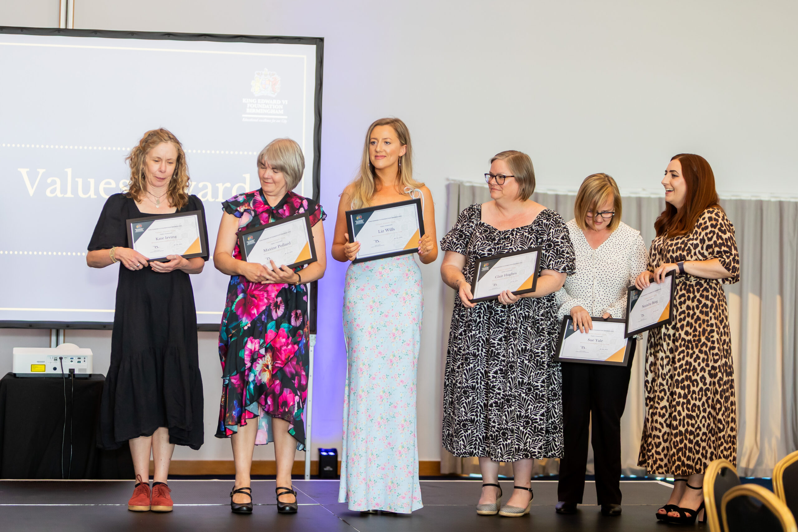 Six women standing on a stage, holding certificates and smiling, likely at an educational award ceremony or event.