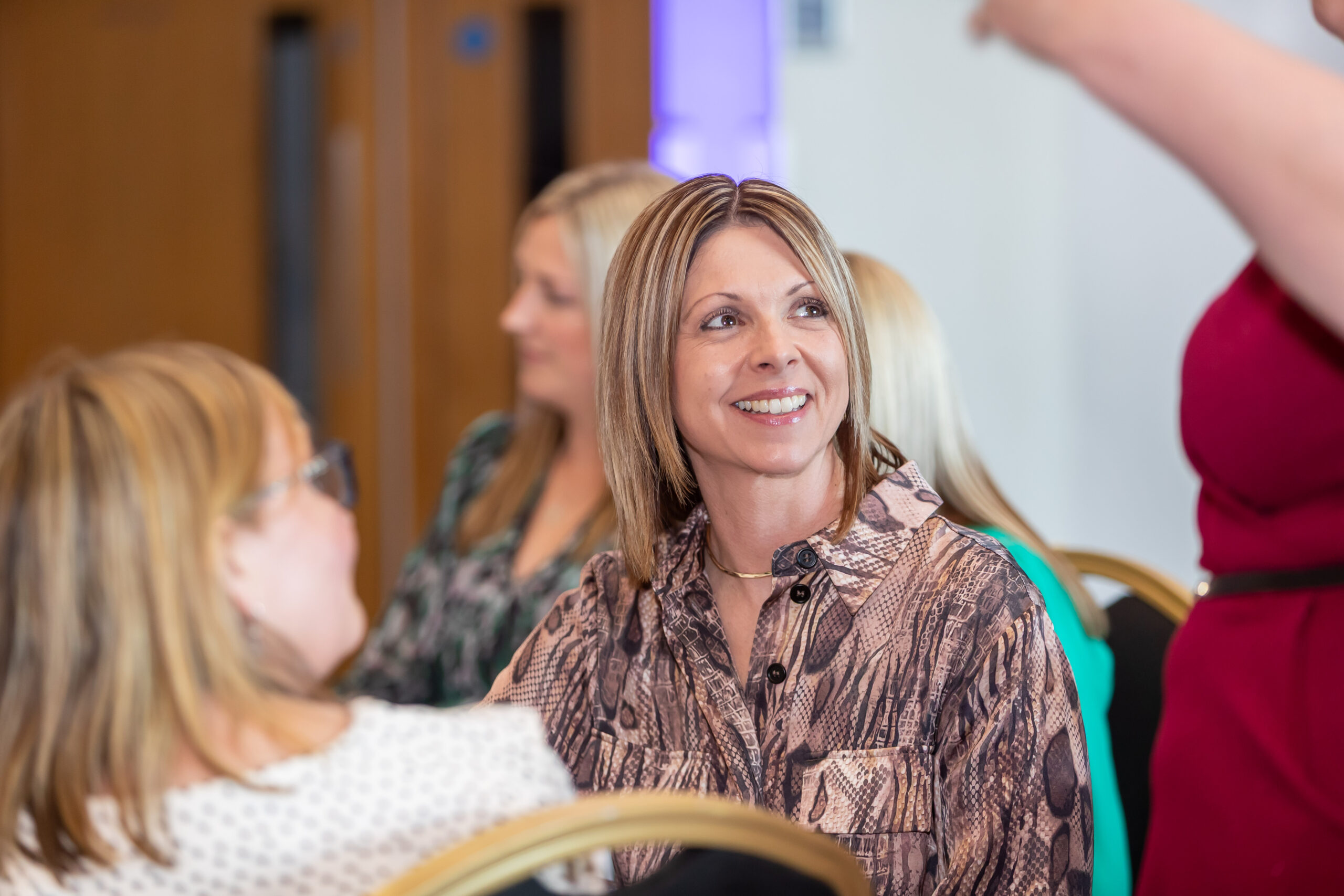 A woman with short blonde hair and a brown patterned shirt smiles while engaging with others in a meeting or workshop setting.