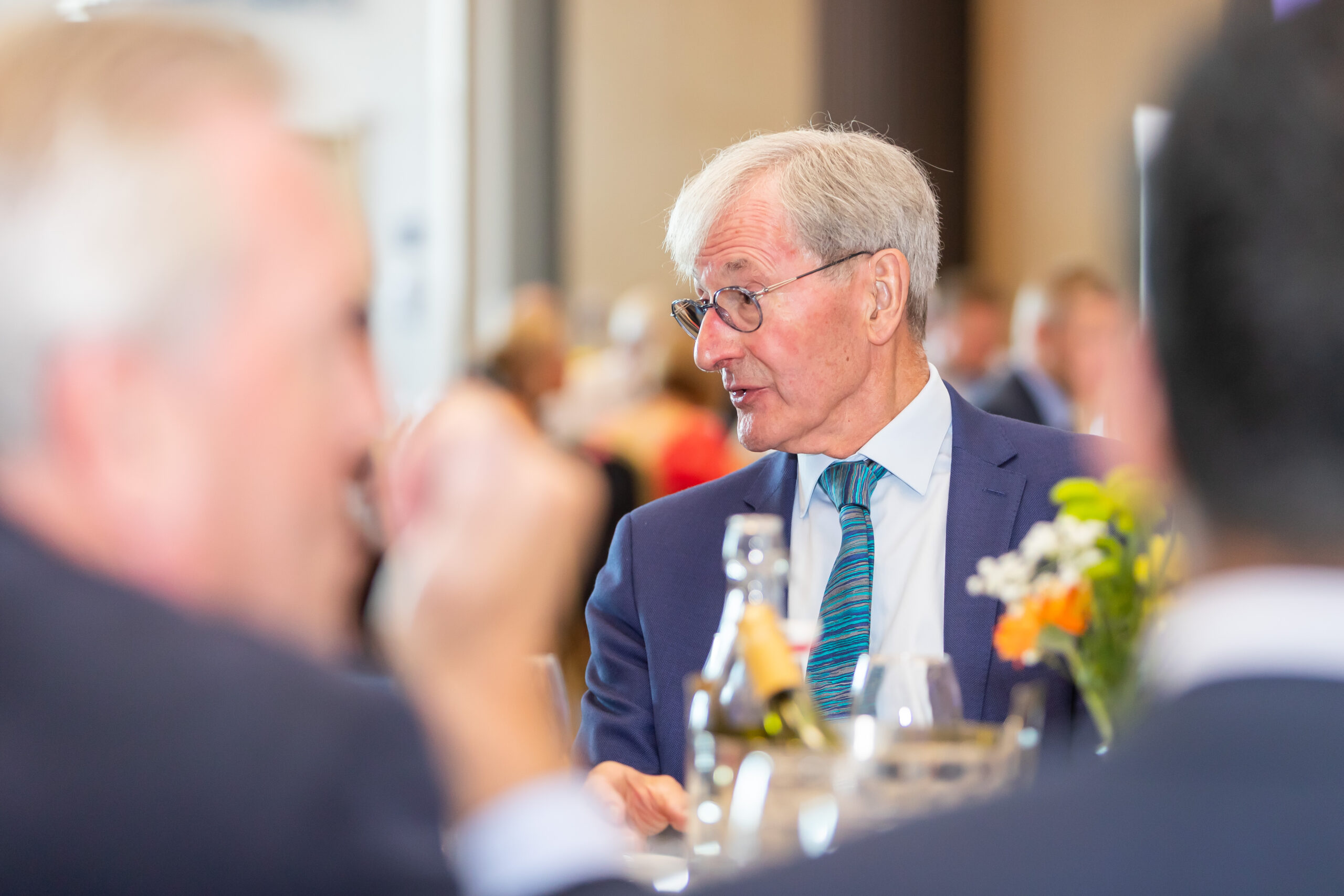 Man in a suit smiling while talking to people at a table with glasses and flowers.