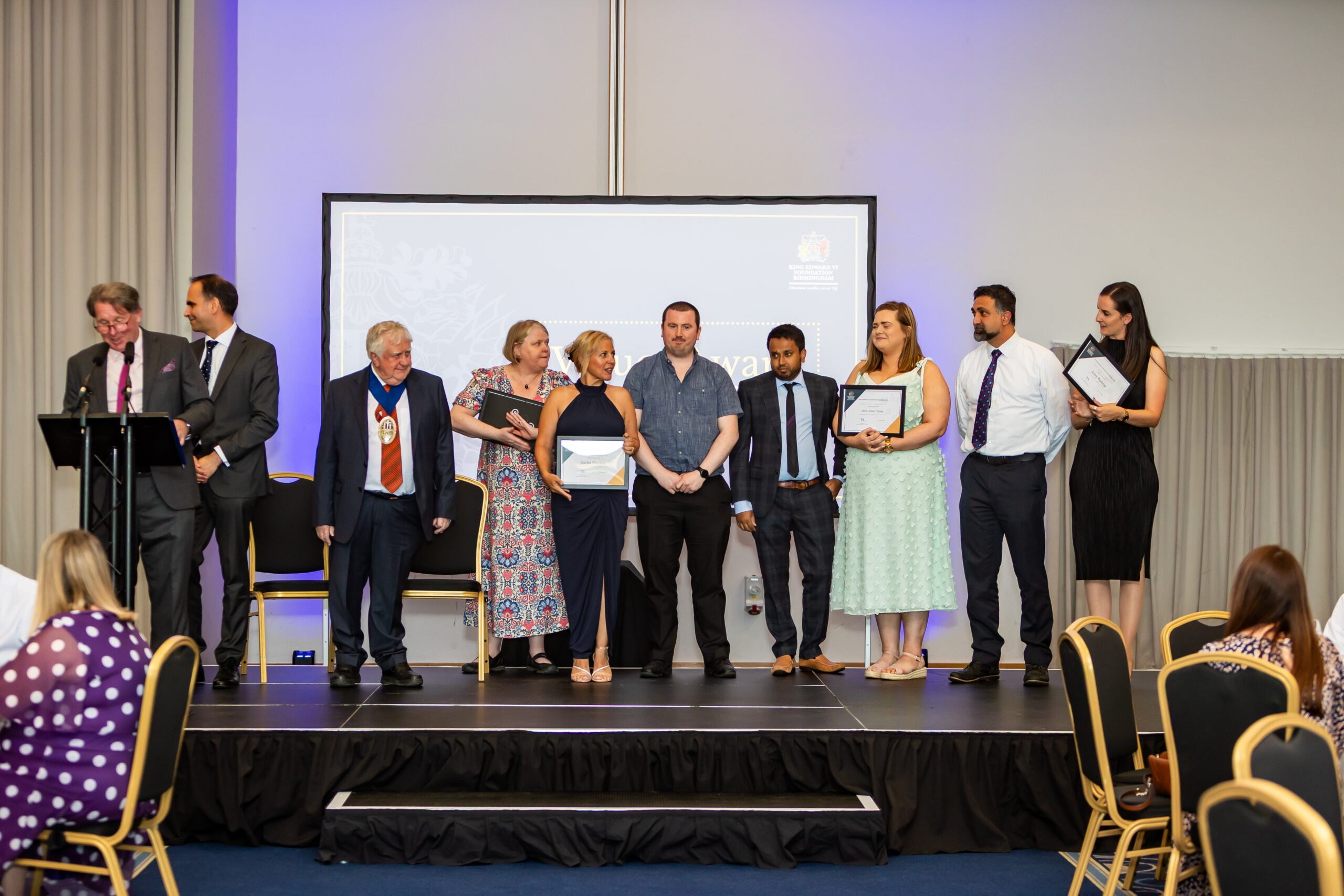 A group of formally dressed people stand on a stage, holding certificates and smiling, with a projector screen behind them.