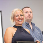 A woman in a black dress smiles while holding an award certificate, flanked by two men in a professional setting.