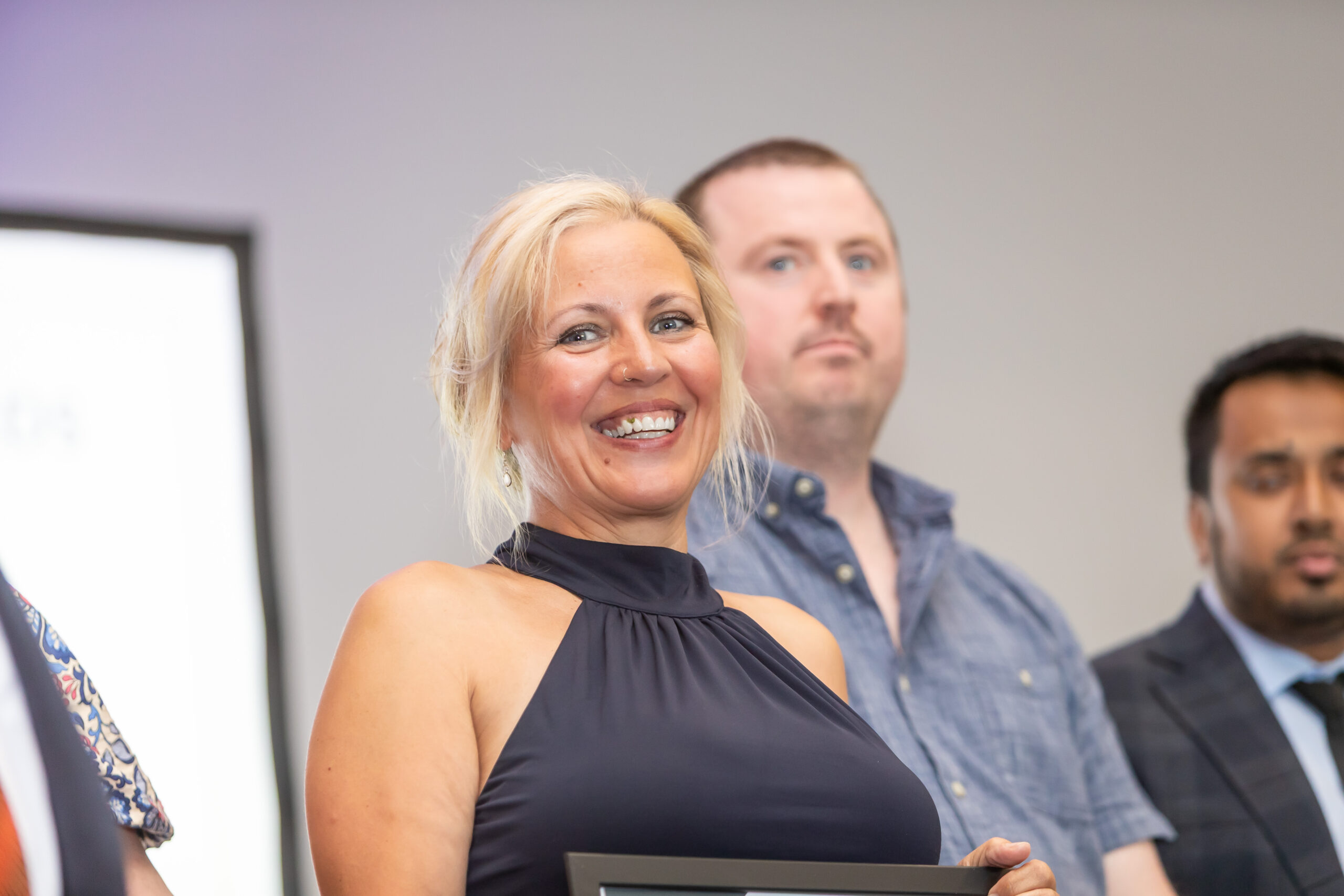 A woman in a black dress smiles while holding an award certificate, flanked by two men in a professional setting.