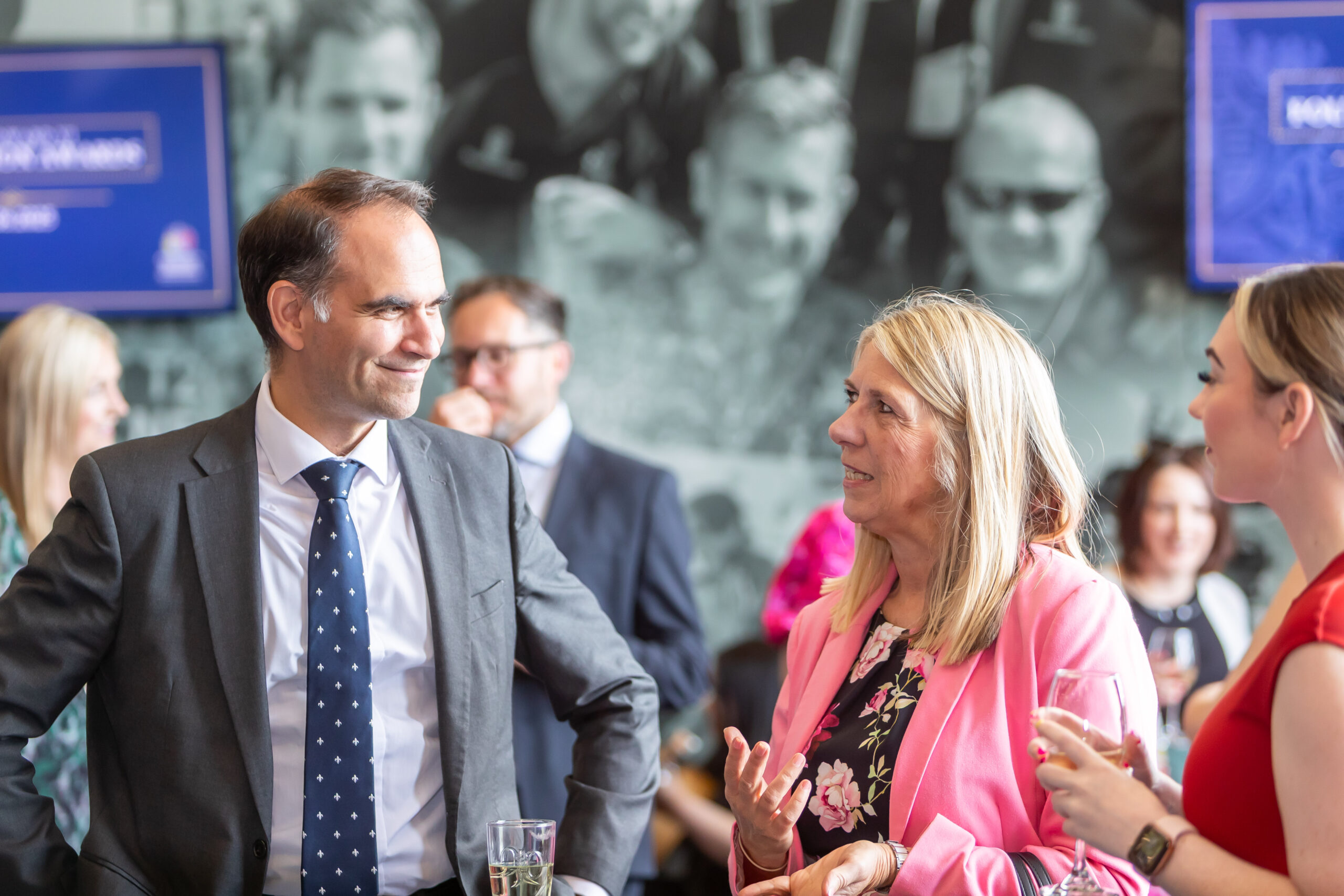 Man and woman in business attire talking at a networking event, with other professionals in the background.
