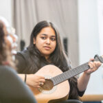 A student plays an acoustic guitar while being observed by another student in a classroom setting, likely during a music lesson or educational activity.