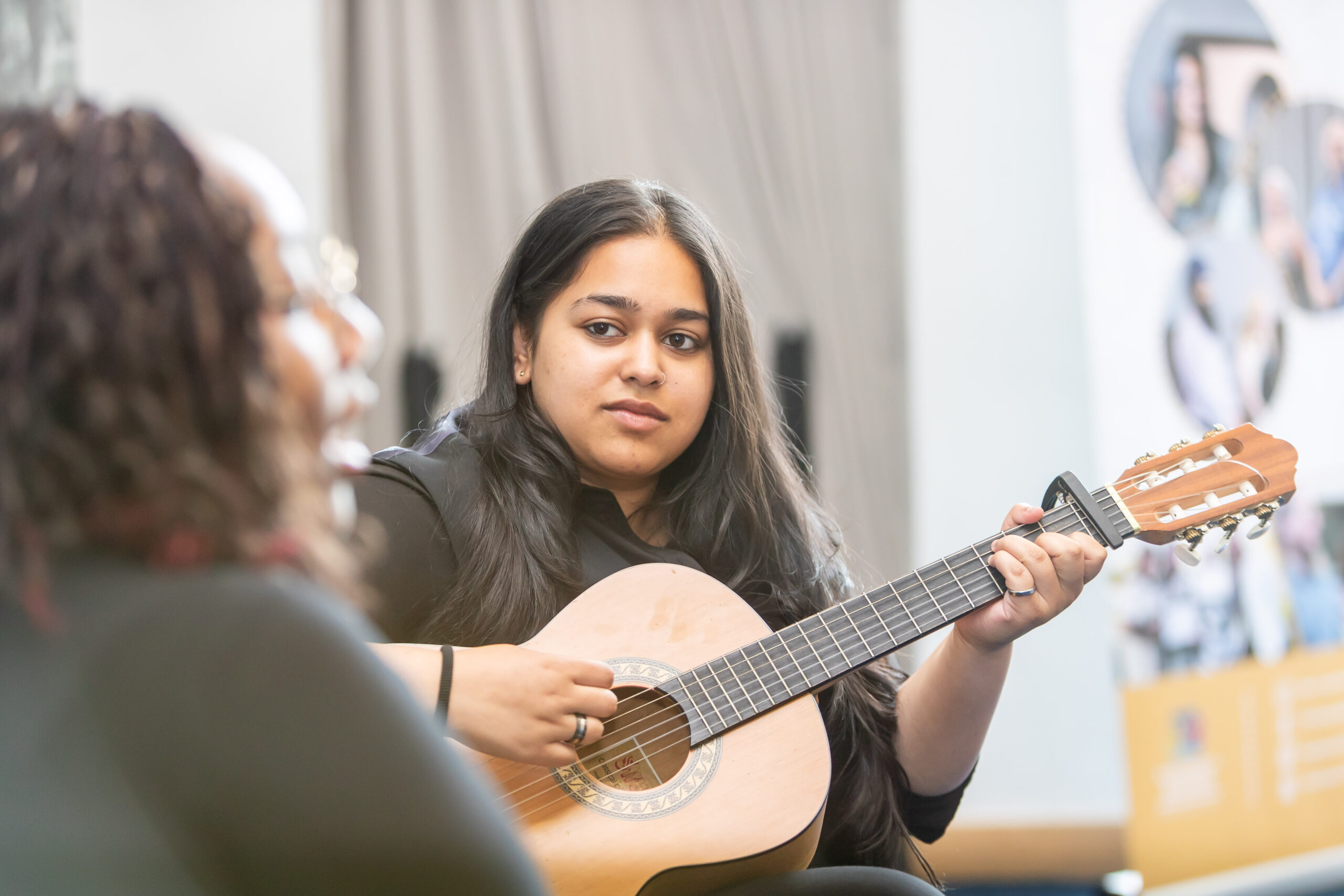 A student plays an acoustic guitar while being observed by another student in a classroom setting, likely during a music lesson or educational activity.
