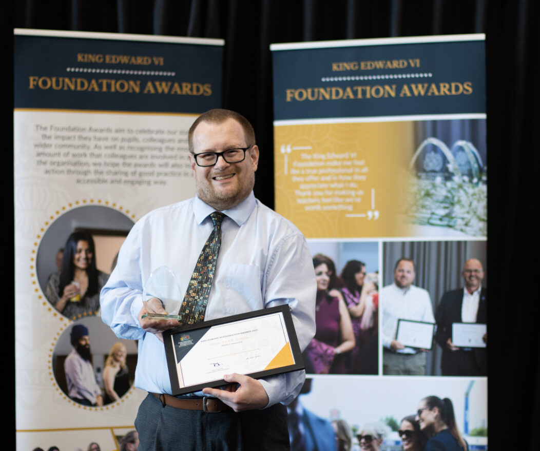 Man holding an award certificate standing in front of Kevi Foundation Awards banners, celebrating educational achievement.