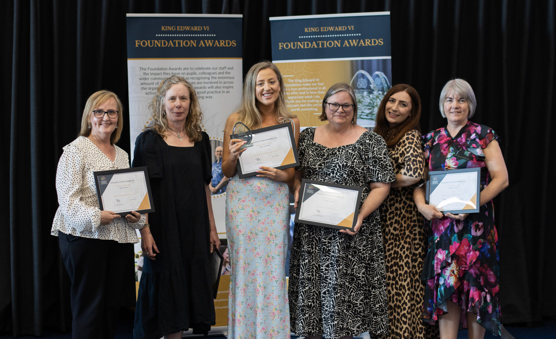 Six women proudly holding certificates on stage at a Kevi Foundation event, celebrating educational achievements.