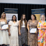 Group of students and teacher holding certificates on stage, posing for a photo with banners in the background, celebrating academic achievement.