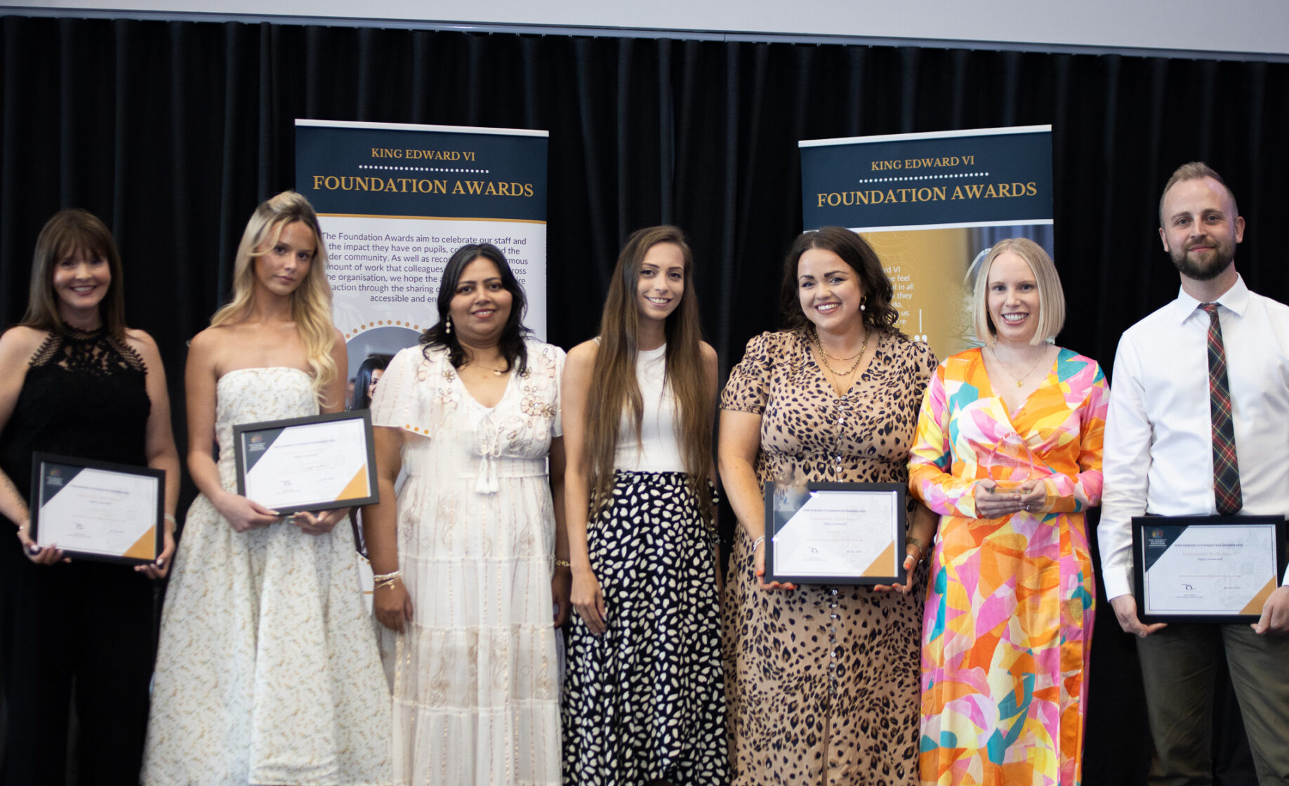 Group of students and teacher holding certificates on stage, posing for a photo with banners in the background, celebrating academic achievement.