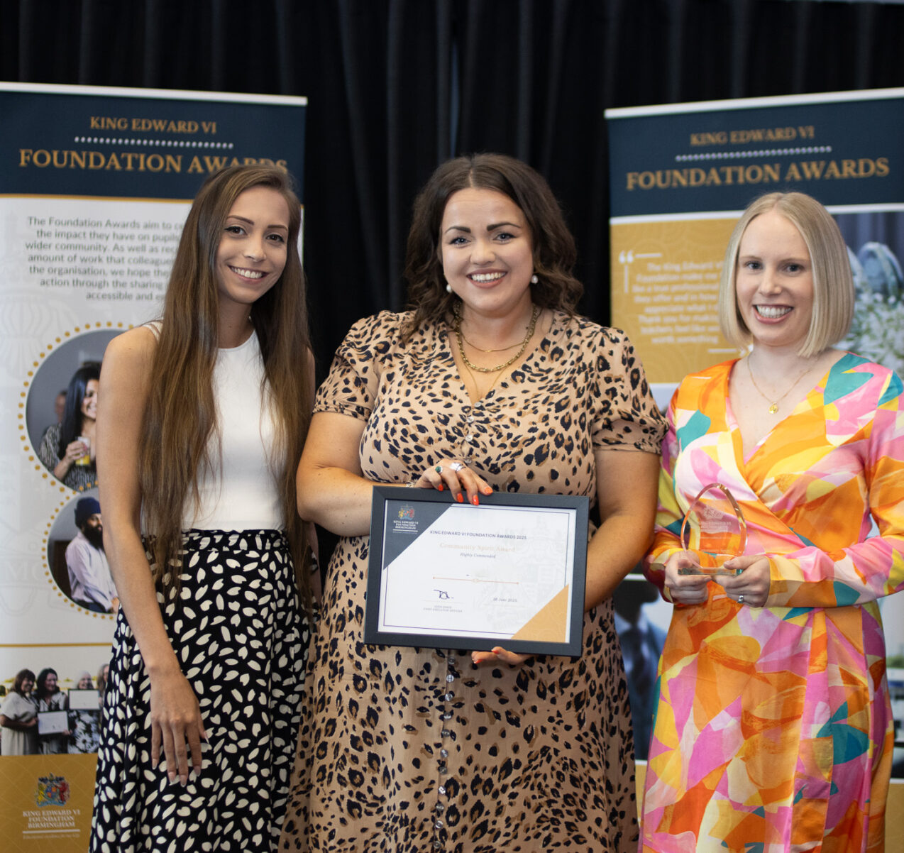 Three women standing together, one holding a certificate, in front of educational banners at the Kevi Foundation event.
