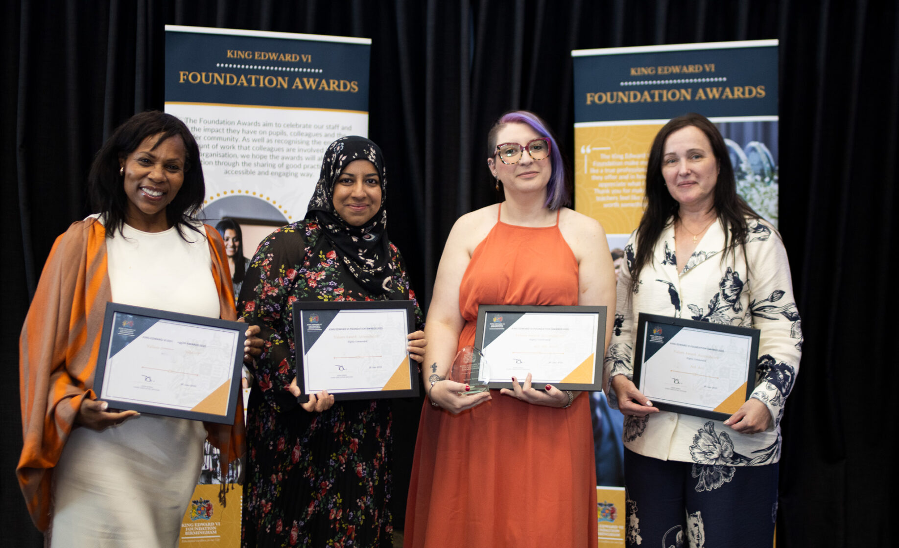 Four women proudly holding certificates in front of a black curtain and Foundation Awards banners, celebrating educational achievements.