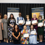 Group of people on a stage proudly holding certificates, smiling and posing for a photo, likely at a graduation or recognition ceremony.