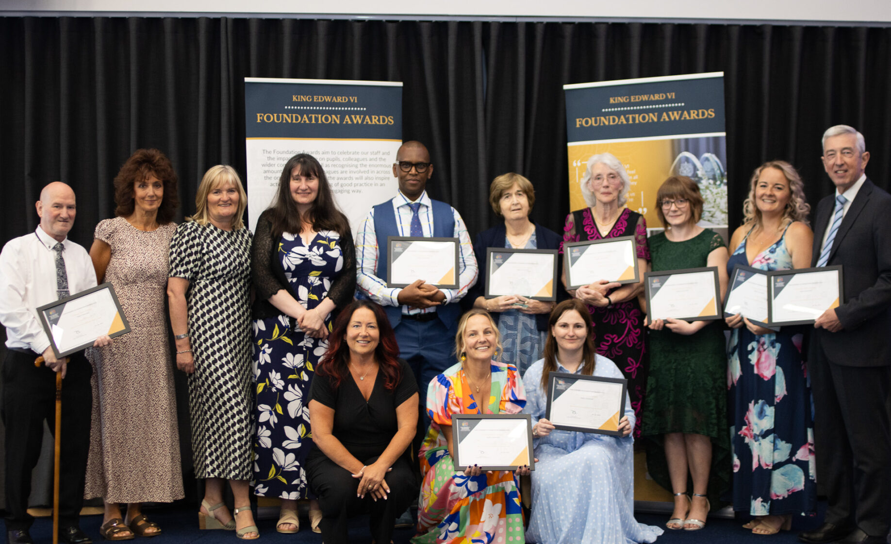 Group of people on a stage proudly holding certificates, smiling and posing for a photo, likely at a graduation or recognition ceremony.