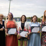 Five women standing on a sports field, smiling and holding certificates, celebrating academic or athletic achievement.