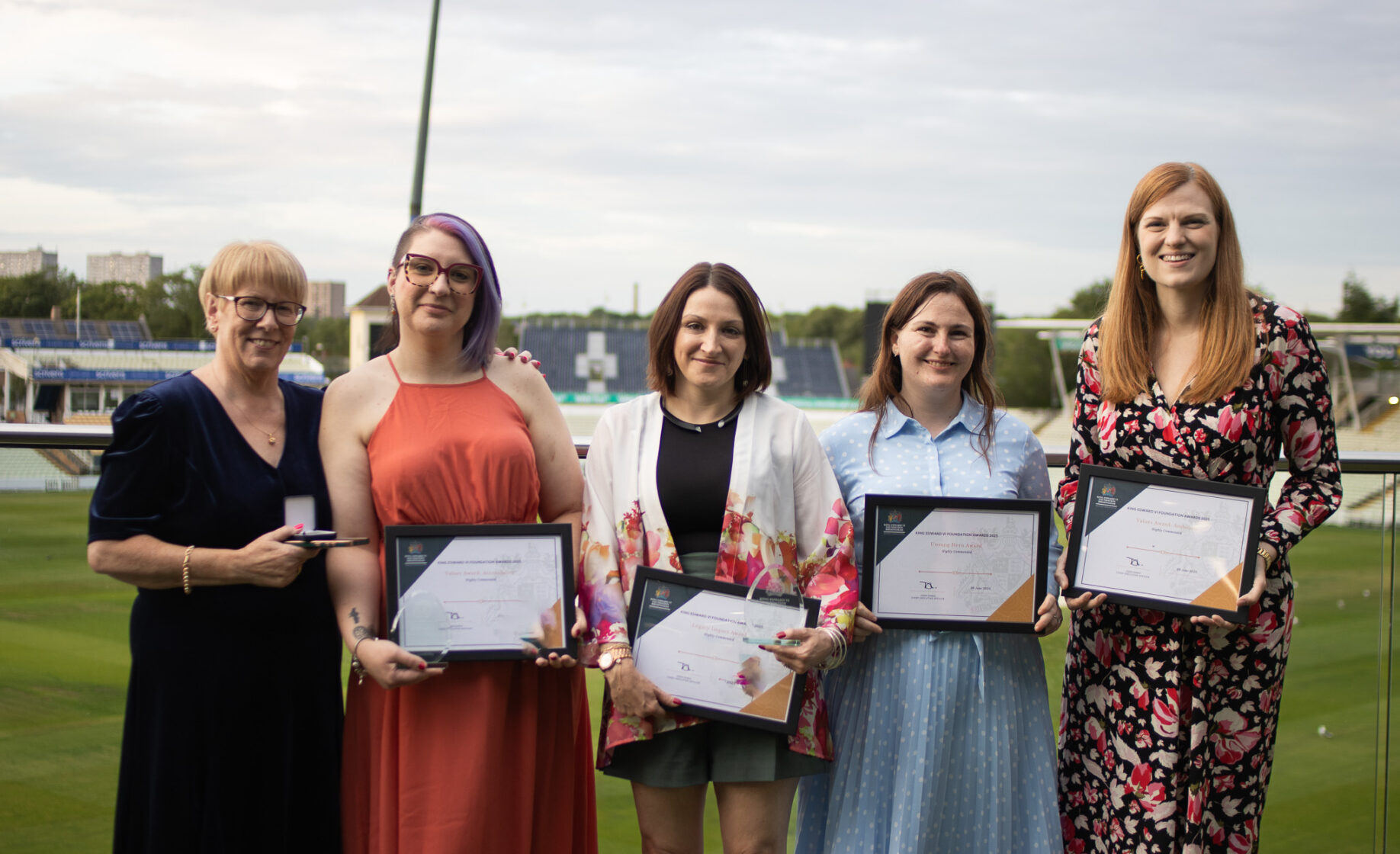 Five women standing on a sports field, smiling and holding certificates, celebrating academic or athletic achievement.
