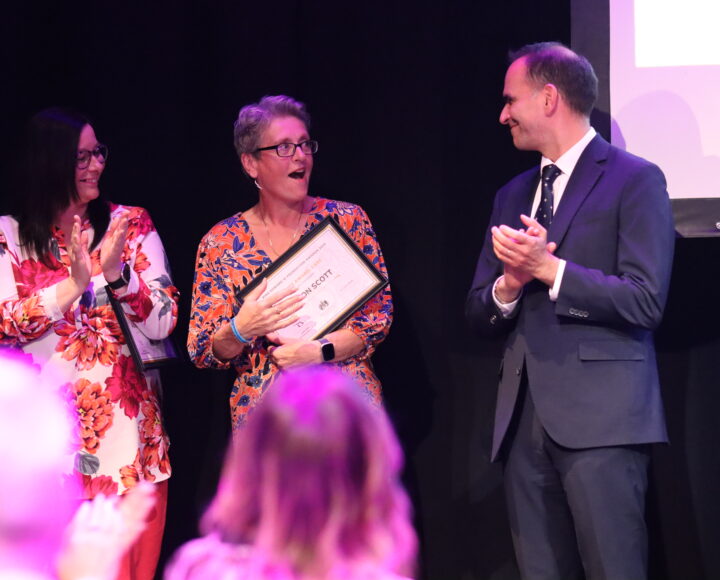 Two women and a man on stage, one woman holding a certificate, at a formal event