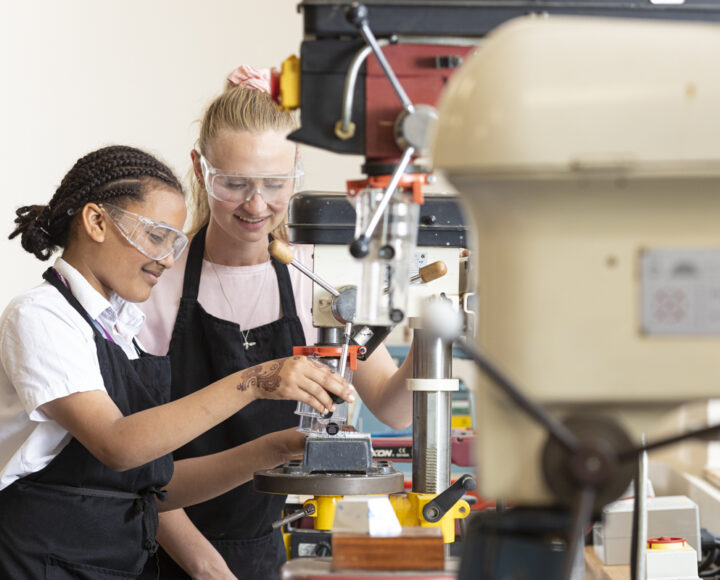 Students using a milling machine in a workshop, wearing safety glasses and black aprons. A close-up of a student operating machinery is visible in the background.